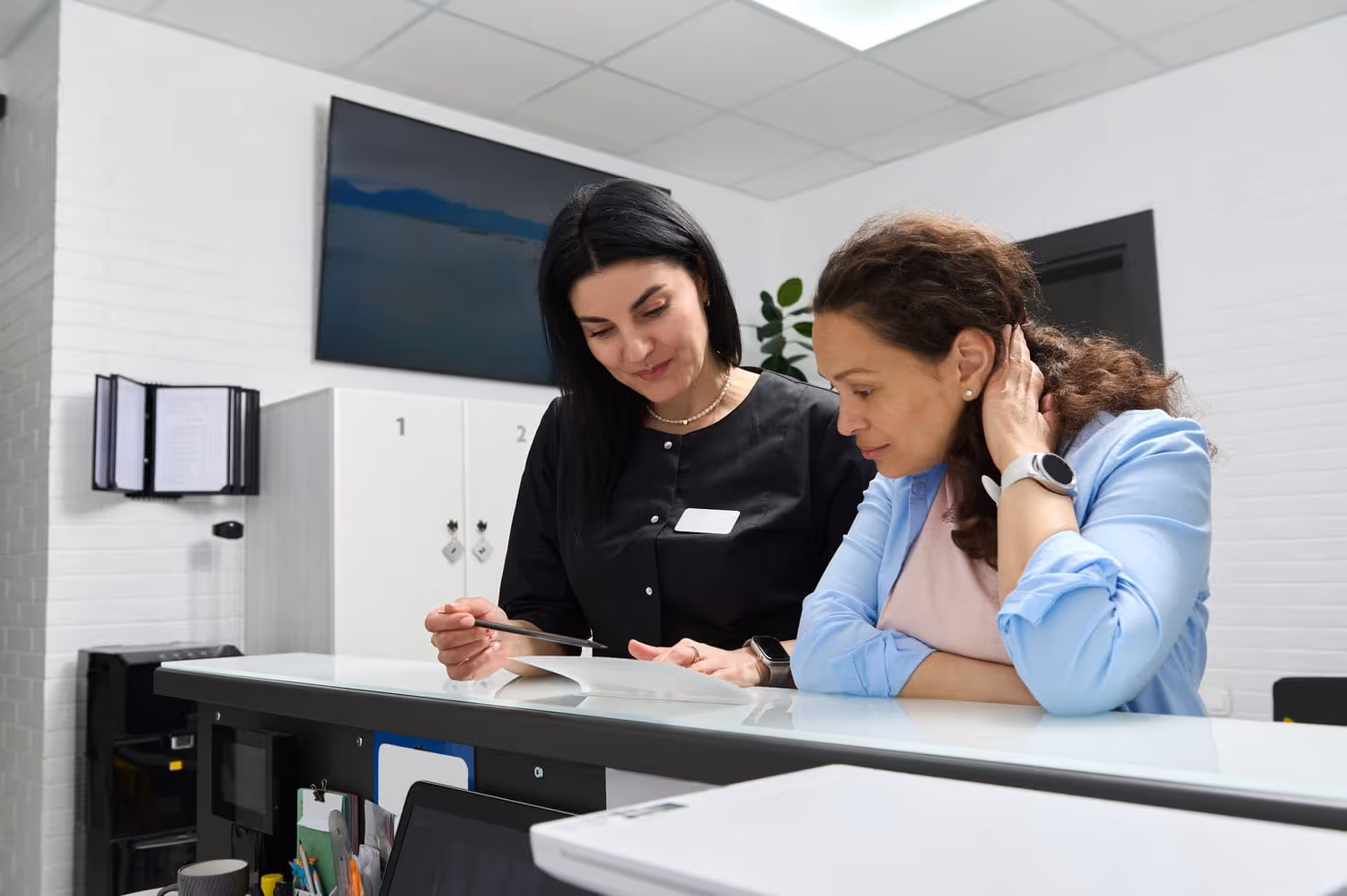 Healthcare receptionist explaining documents to a patient at the front desk in a medical office.